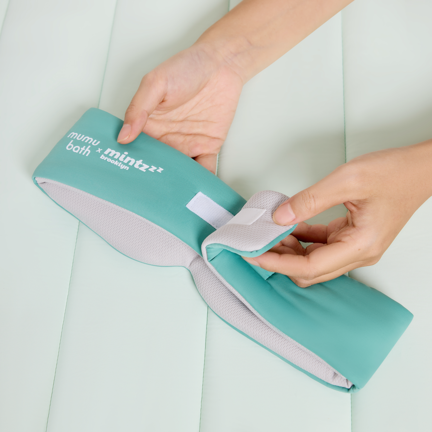 Two hands adjust the velcro strap on a teal and gray Mintzzz "Chill Mask | Cooling Sleep Mask" against a light padded surface.