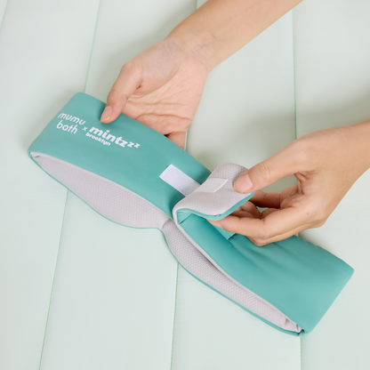 Two hands adjust the velcro strap on a teal and gray Mintzzz "Chill Mask | Cooling Sleep Mask" against a light padded surface.