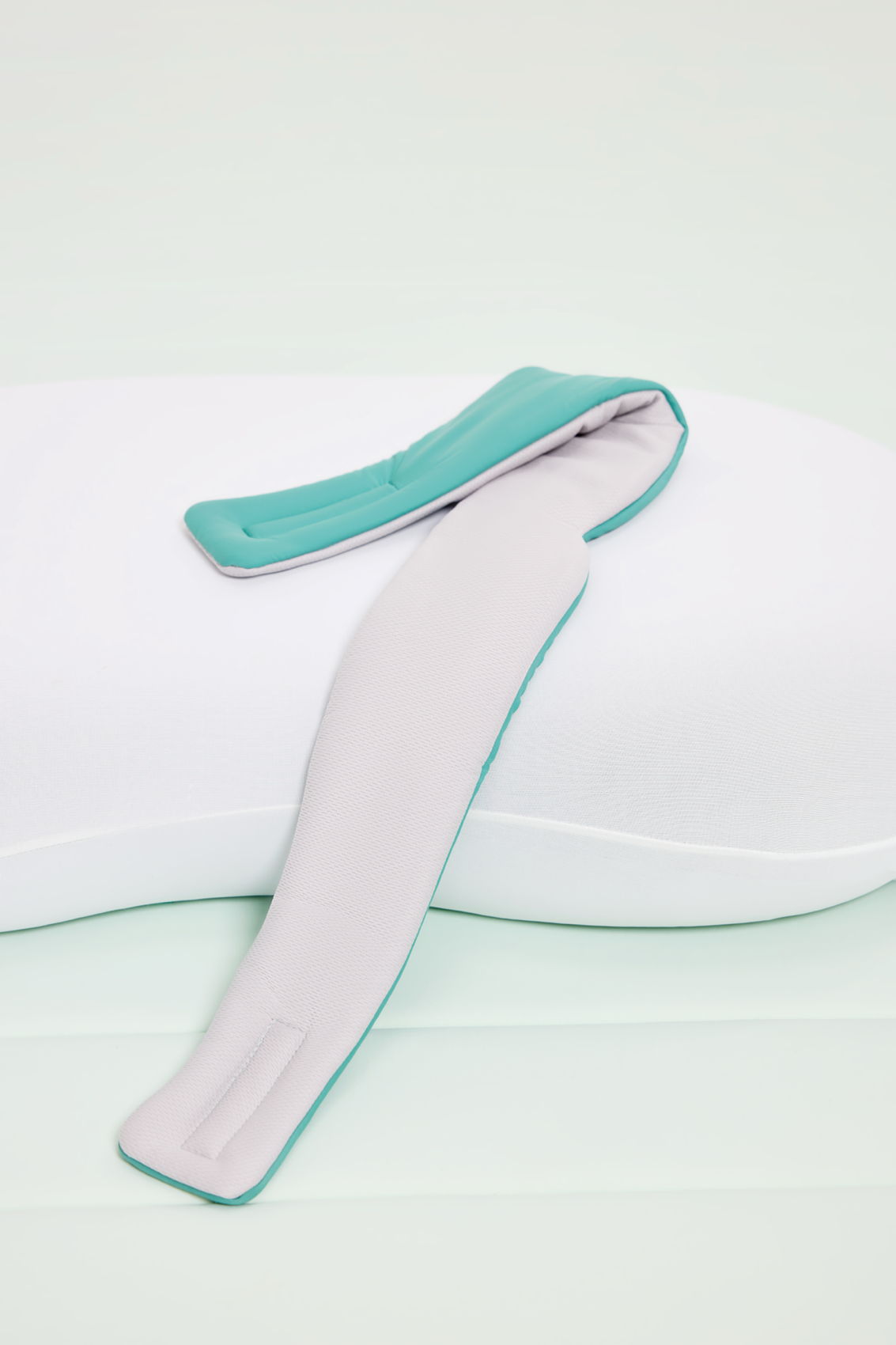 A white ergonomic pillow with a light gray and teal padded strap sits on a light surface—perfectly paired with the Mintzzz Chill Mask | Cooling Sleep Mask for optimal rest.